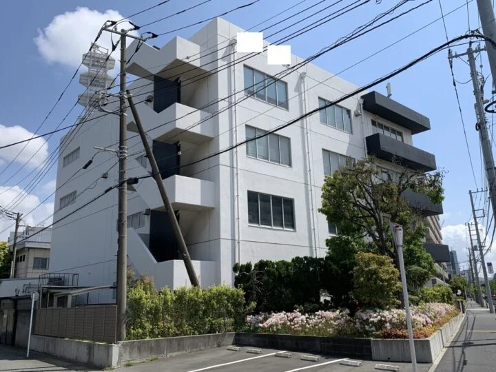 白い外壁と黒いバルコニーが特徴の4階建てマンション外観。道路沿いの角地に建ち、植栽と電柱が周囲に見える施工前の状態